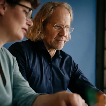 Zwei Personen, eine mit Brille und kurzem Haar, die andere mit schulterlangem, blondem Haar, blicken auf einen Bildschirm, die Person mit Brille gestikuliert mit der Hand.