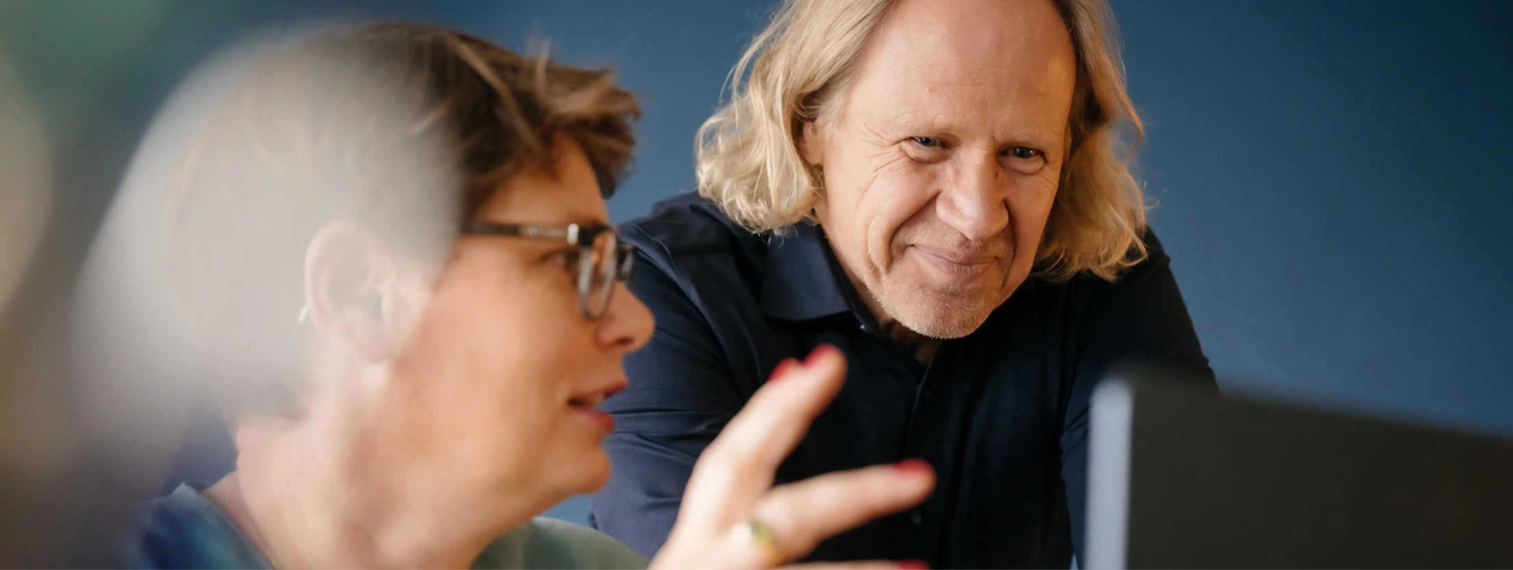 Zwei Personen in einem Gespräch, Petra von Notz mit kurzen Haaren und Brille gestikuliert, Michael Hirth mit schulterlangen blonden Haaren lächelt und schaut auf einen Bildschirm.