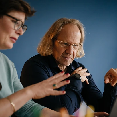 Zwei Personen, eine mit Brille und kurzem Haar, die andere mit schulterlangem, blondem Haar, blicken auf einen Bildschirm, die Person mit Brille gestikuliert mit der Hand.