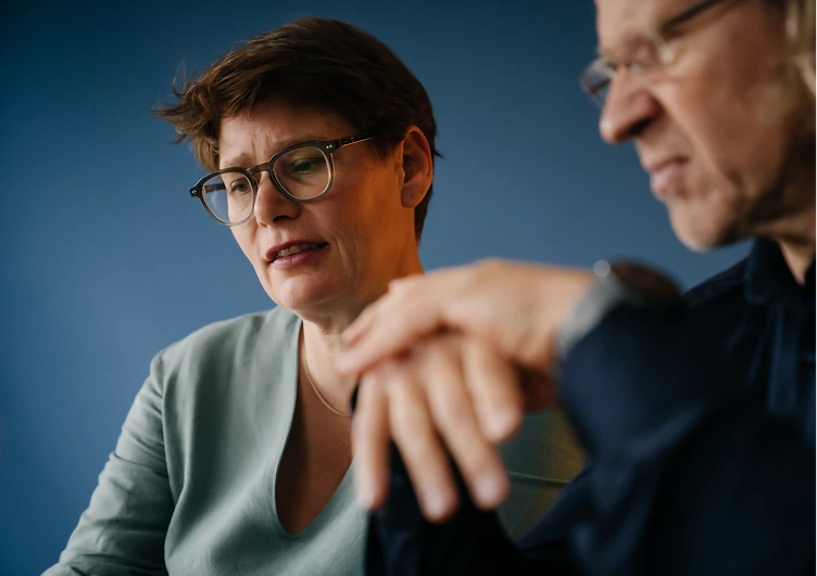 Zwei Personen in Nahaufnahme vor blauem Hintergrund, Petra von Notz mit Brille und kurz geschnittenem Haar blickt konzentriert, Michael Hirth mit Brille und grauem Haar ist unscharf im Vordergrund zu sehen.